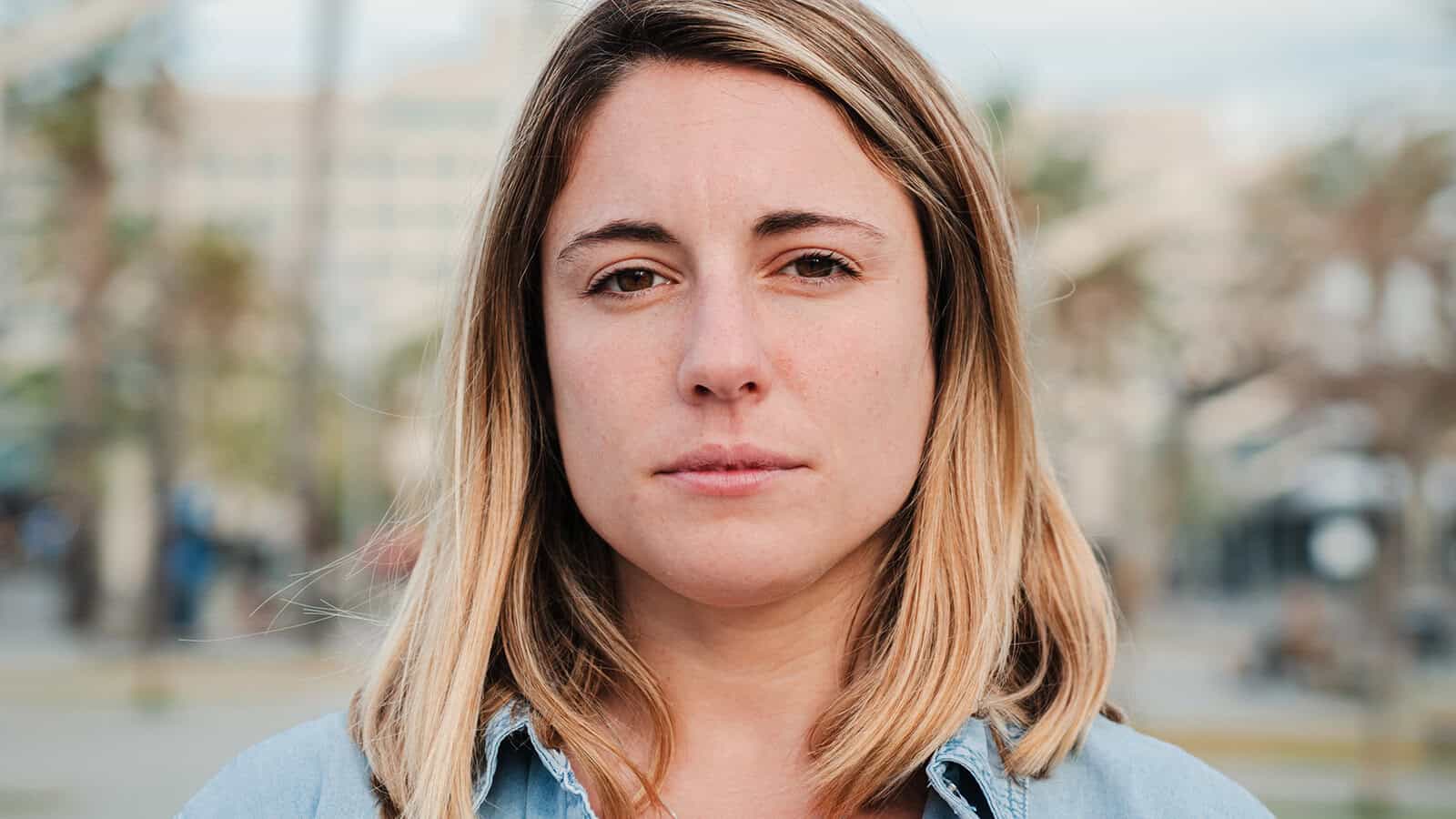 A young woman with blonde hair and a neutral expression looks directly at the camera. She is outdoors, with blurred trees and buildings in the background.