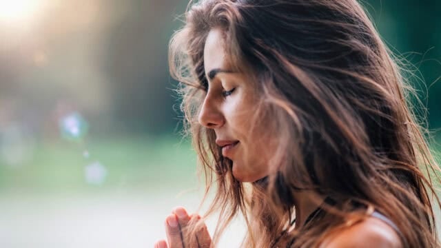 A woman with long brown hair stands outdoors with her eyes closed, hands together in a prayer position, appearing peaceful and reflective, with sunlight filtering through the background.