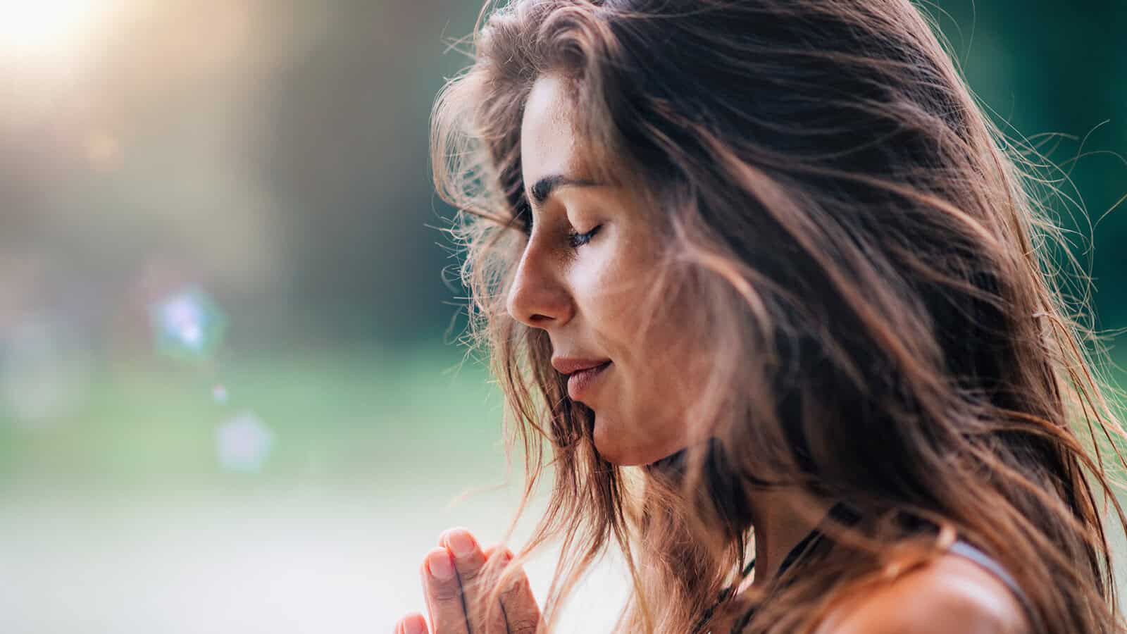 A woman with long brown hair stands outdoors with her eyes closed, hands together in a prayer position, appearing peaceful and reflective, with sunlight filtering through the background.