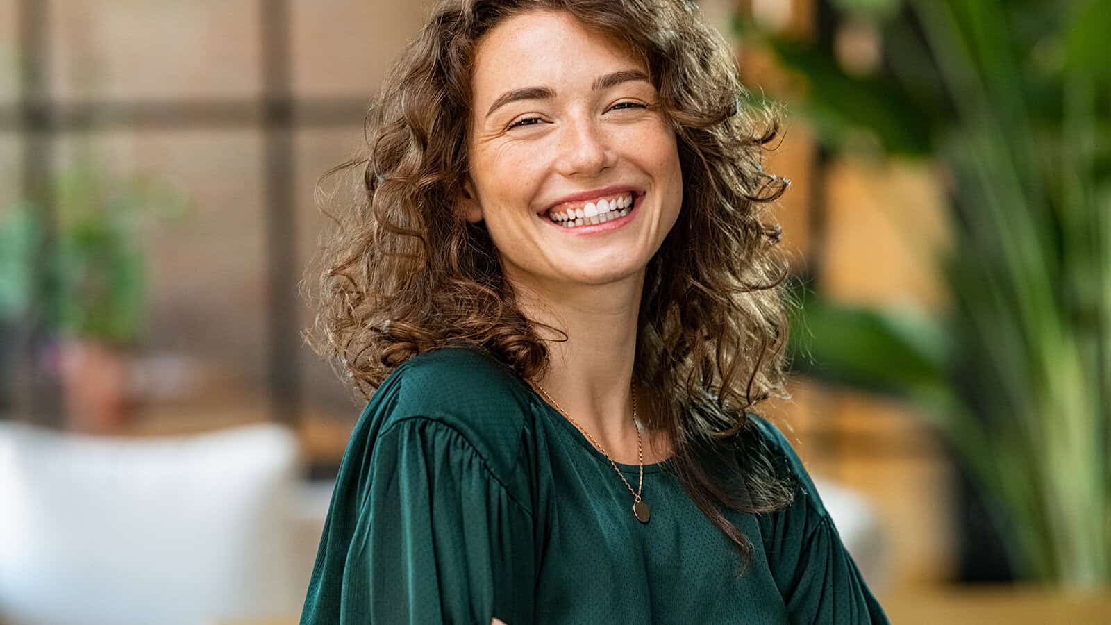 Smiling woman with curly brown hair wearing a dark green blouse, standing indoors with blurred plants and windows in the background.