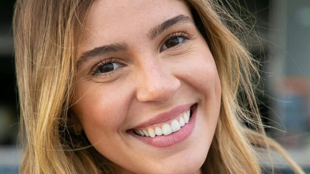 A young woman with long, light brown hair and brown eyes smiles warmly at the camera, showing her teeth. The background is blurred.
