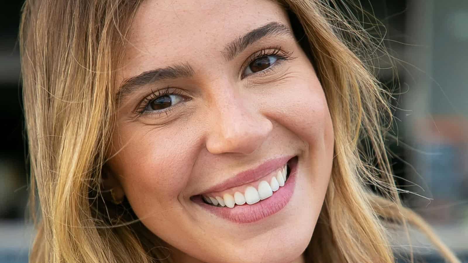 A young woman with long, light brown hair and brown eyes smiles warmly at the camera, showing her teeth. The background is blurred.