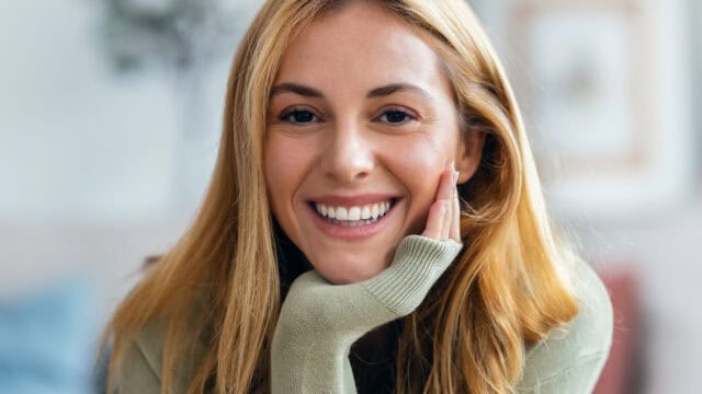 A young woman with long blonde hair, exuding emotional maturity, smiles warmly at the camera, resting her chin on her hand. She wears a light green sweater and is indoors, with a blurred background.