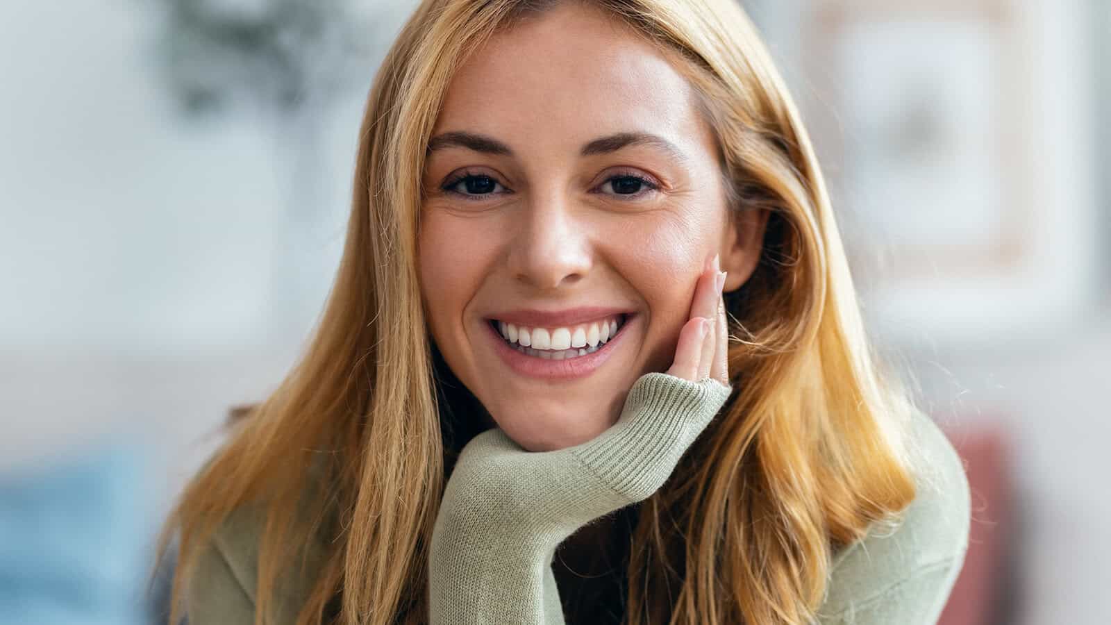 A young woman with long blonde hair, exuding emotional maturity, smiles warmly at the camera, resting her chin on her hand. She wears a light green sweater and is indoors, with a blurred background.