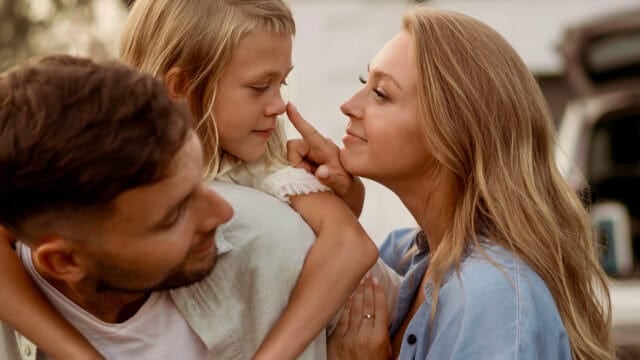 A woman gently touches a young girls nose while a man holds the girl; all three are smiling and sharing a tender, close moment outdoors.