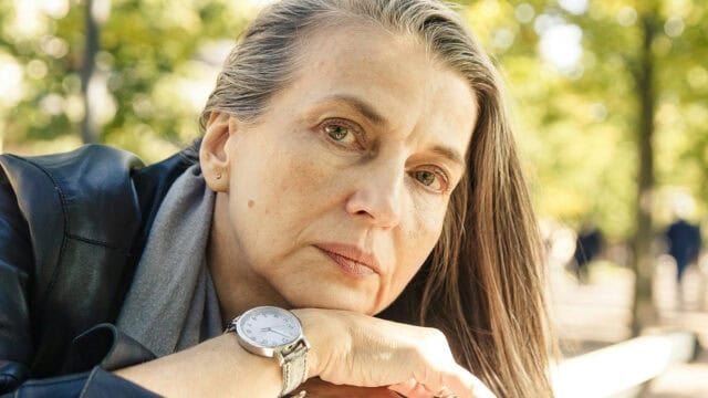 A woman with long gray hair and green eyes leans her head on her crossed arms outdoors, wearing a black jacket, gray scarf, and a silver watch, with a blurred background of trees and sunlight.