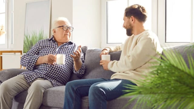 An older man and a younger man sit on a couch, holding mugs and having a conversation in a bright, sunlit room with green plants and large windows in the background.