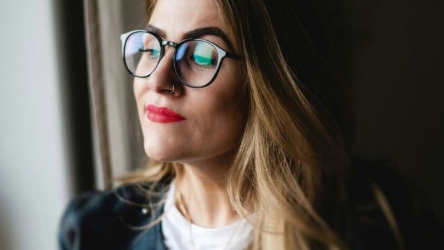 Woman with long blonde hair, wearing glasses and red lipstick, looks out a window with a calm expression.
