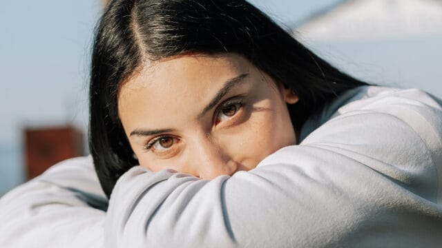 A person with long dark hair rests their chin on their arms, looking directly at the camera with brown eyes. They are wearing a light-colored top and are outdoors in natural light.