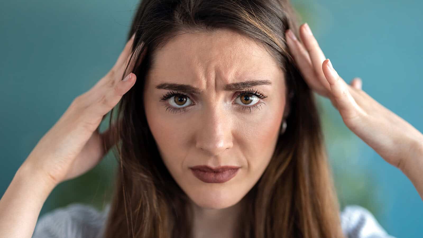 A woman with long brown hair looks worried, holding her hands to her head with a furrowed brow and serious expression, against a blurred blue background.