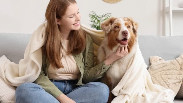 A woman sits on a couch smiling at a dog next to her. Both are covered with a light-colored blanket. The setting appears cozy and homey with neutral decor in the background.