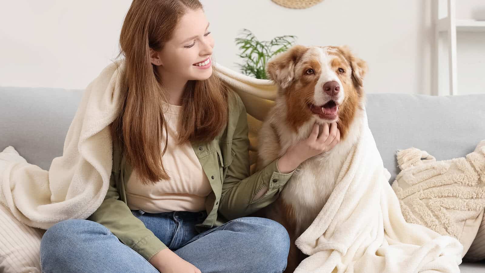 A woman sits on a couch smiling at a dog next to her. Both are covered with a light-colored blanket. The setting appears cozy and homey with neutral decor in the background.
