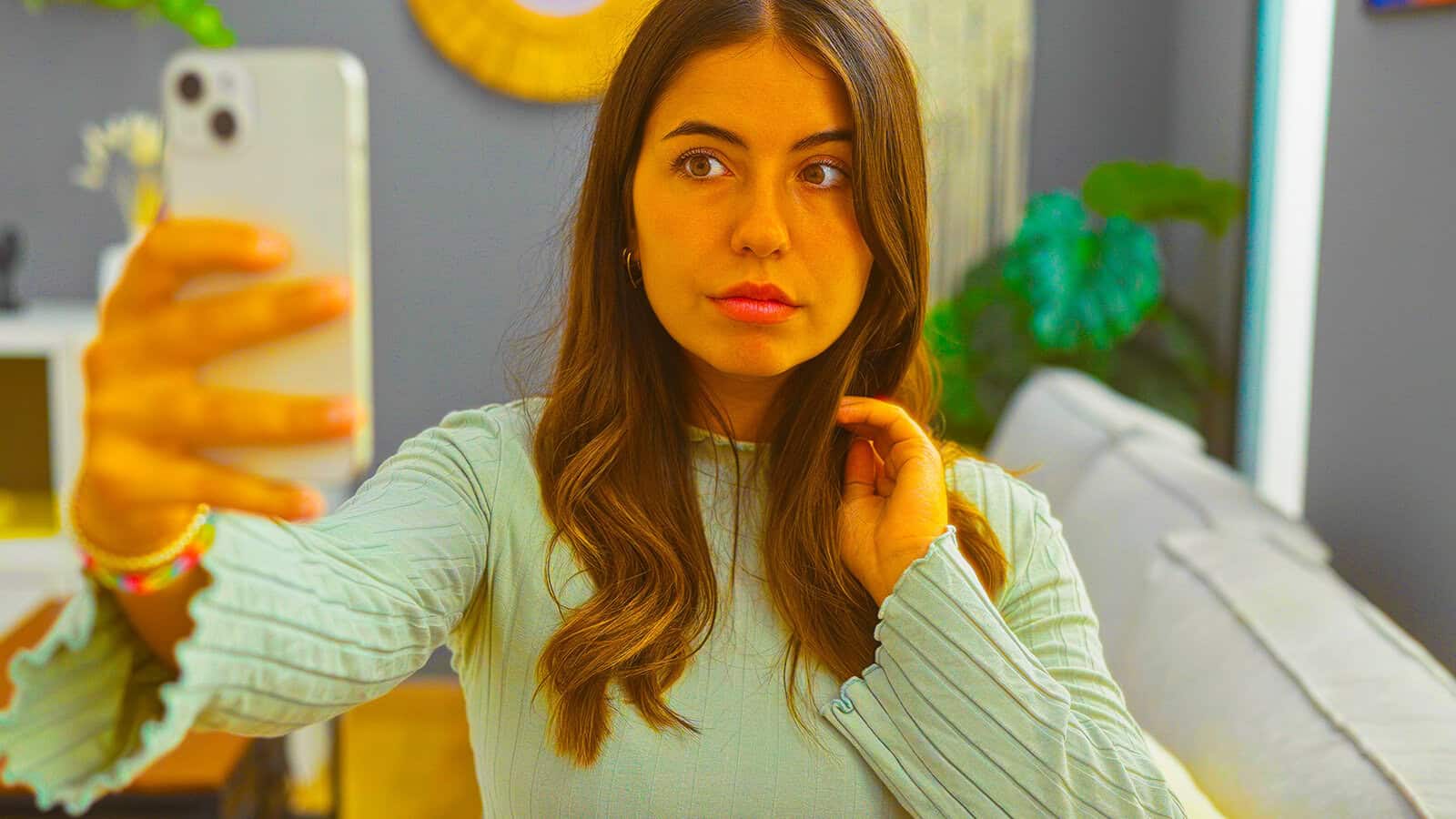A young woman with long brown hair takes a selfie indoors, wearing a light blue top. She holds her phone with one hand and touches her hair with the other. A couch and plants are visible in the background.