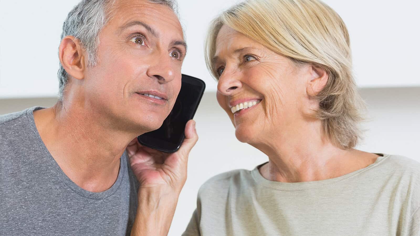 An older man holds a smartphone to his ear, looking curious, while an older woman beside him smiles warmly, both appearing engaged in a conversation.