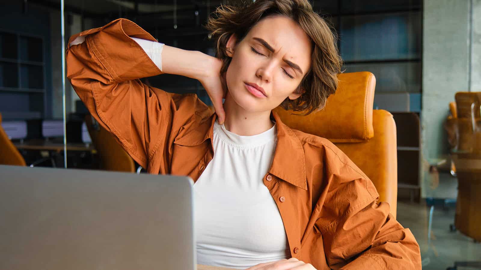 A person sitting at a desk with a laptop, eyes closed and hand on their neck, appears to be experiencing neck pain or discomfort.