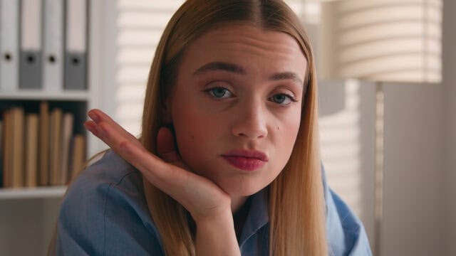 A young woman with long blonde hair rests her chin on her hand, looking directly at the camera with a neutral expression. She is indoors, with bookshelves and a lamp in the background.