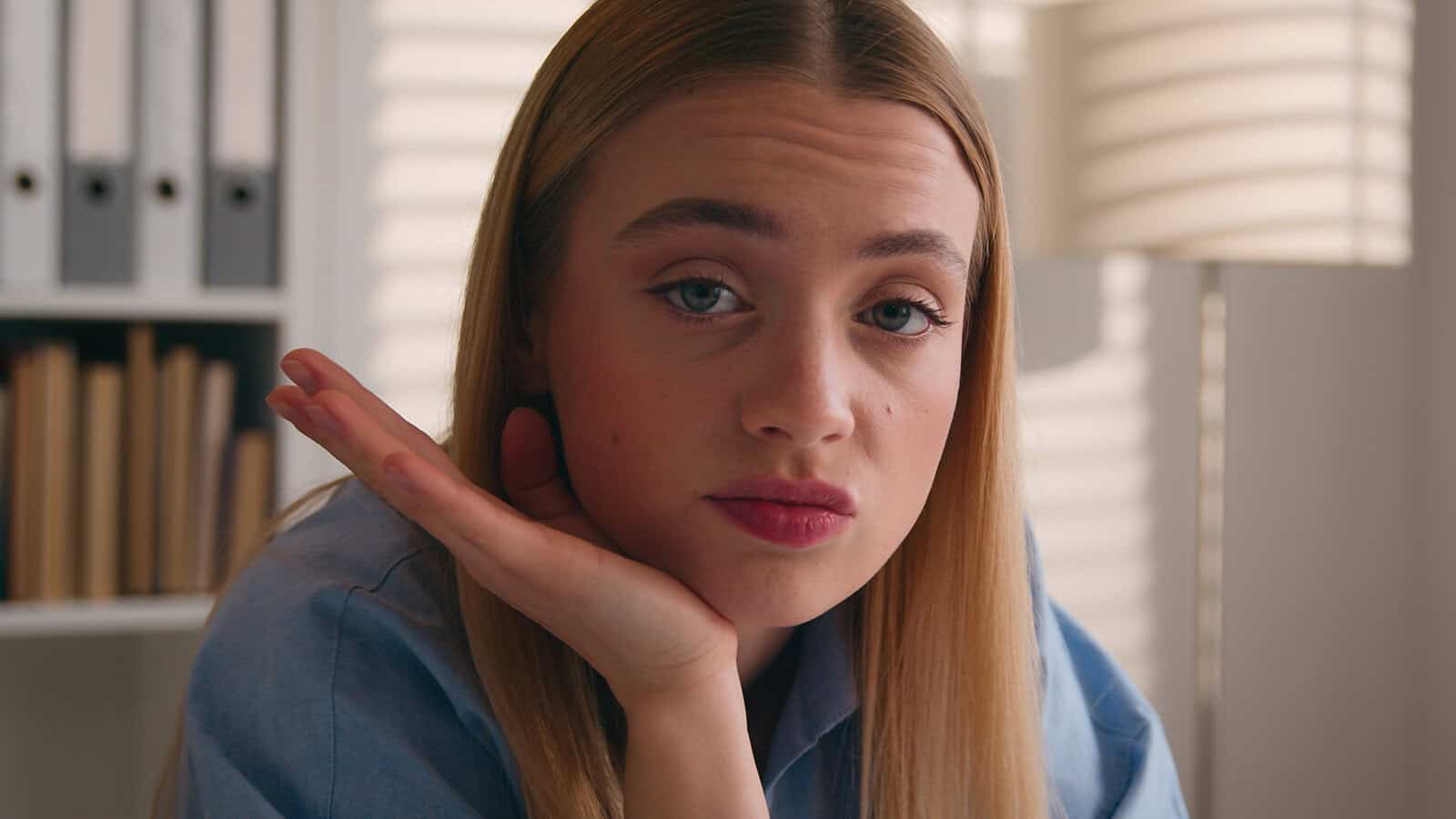 A young woman with long blonde hair rests her chin on her hand, looking directly at the camera with a neutral expression. She is indoors, with bookshelves and a lamp in the background.