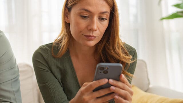 A woman with long hair in a green top sits on a couch, looking intently at her smartphone.