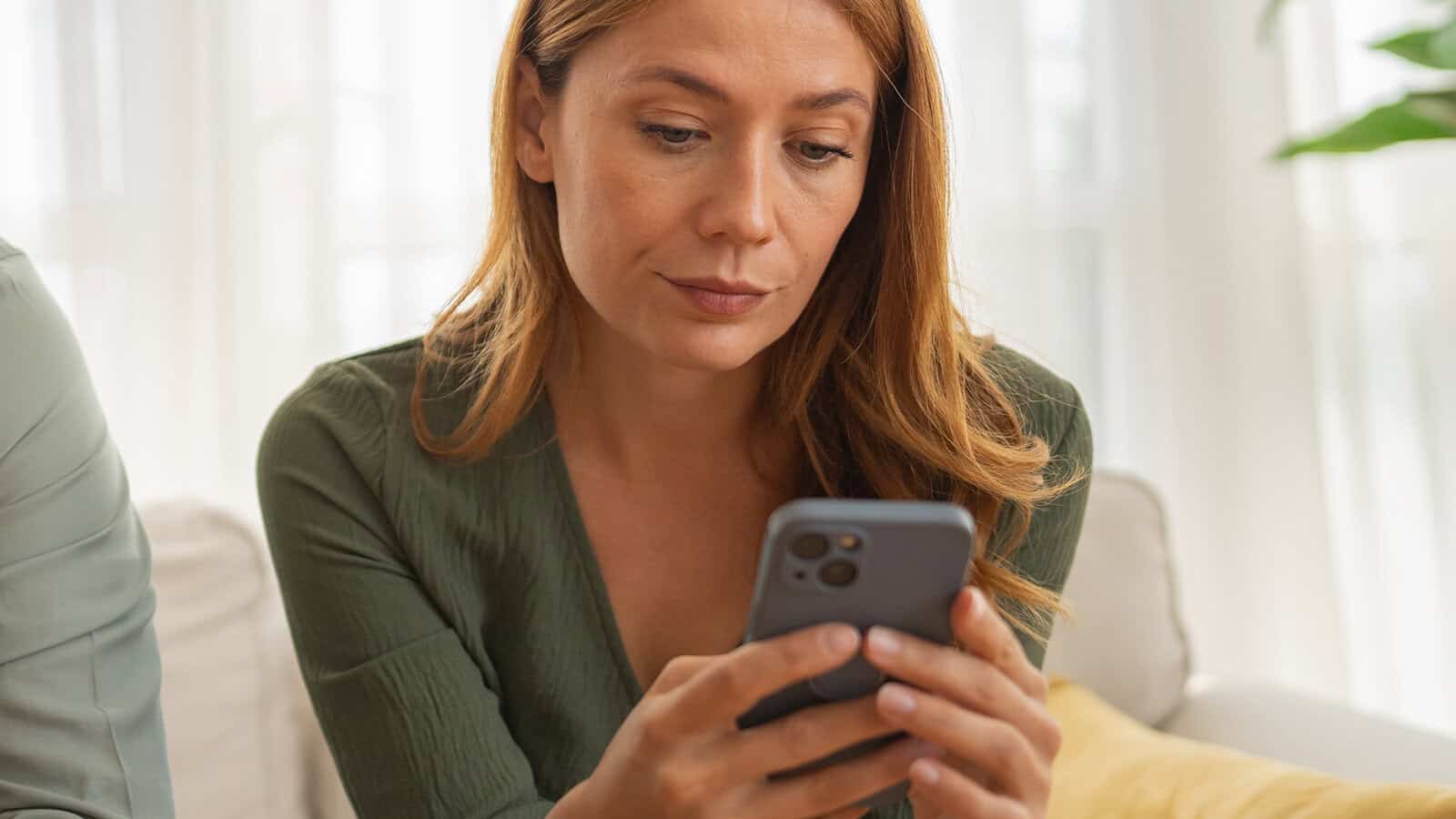A woman with long hair in a green top sits on a couch, looking intently at her smartphone.