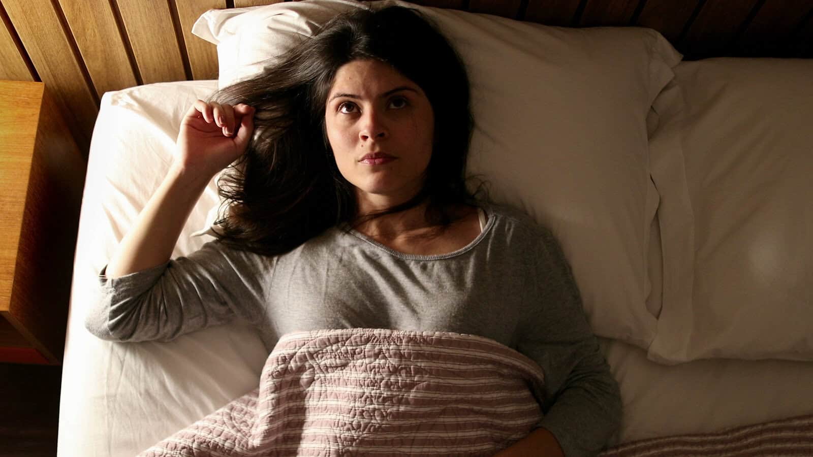 A woman with long dark hair lies in bed under a striped blanket, staring upward with a serious, thoughtful expression, in a dimly lit room.