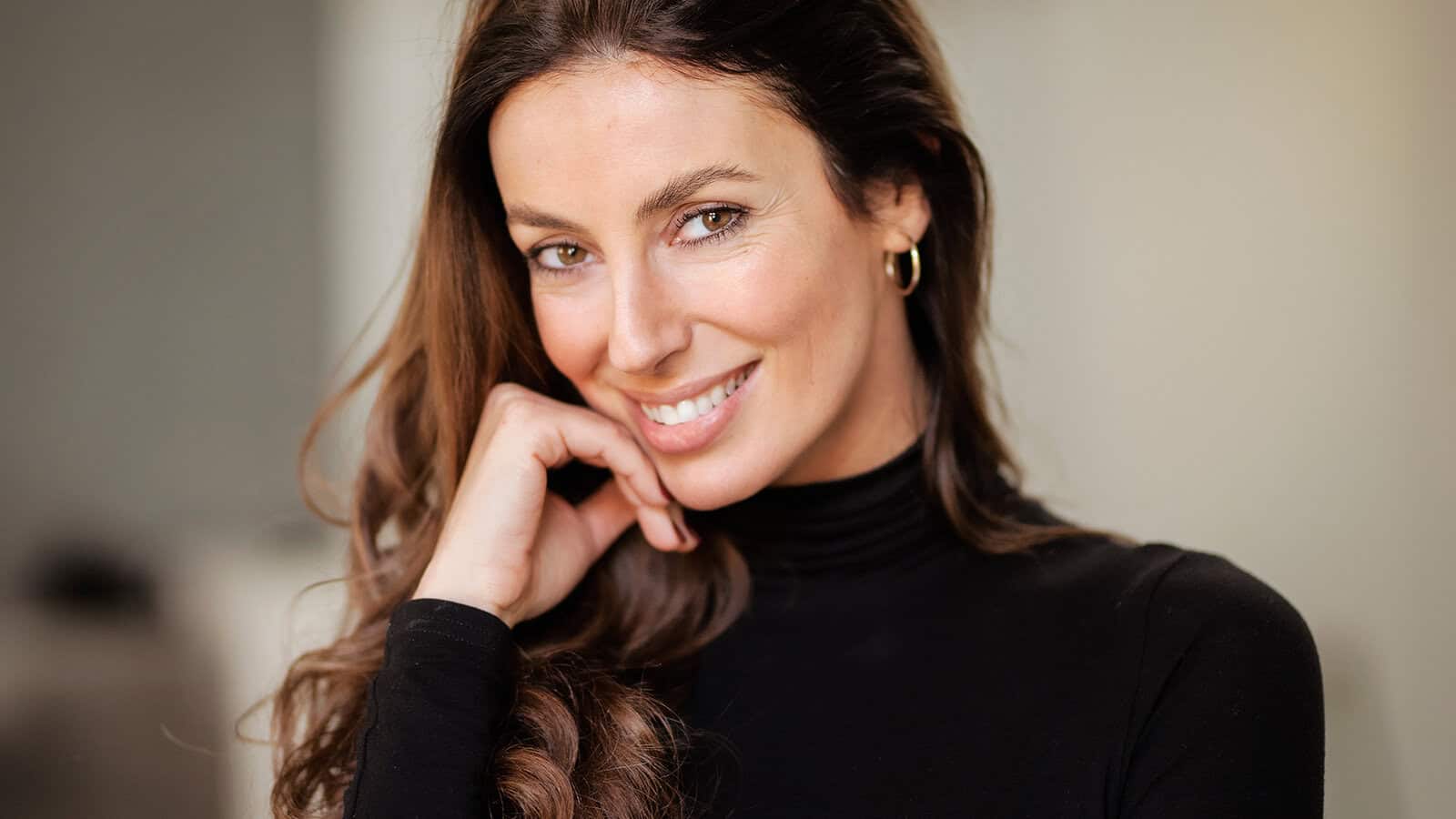 A woman with long brown hair, wearing a black turtleneck and gold hoop earring, smiles while resting her chin on her hand against a blurred background.