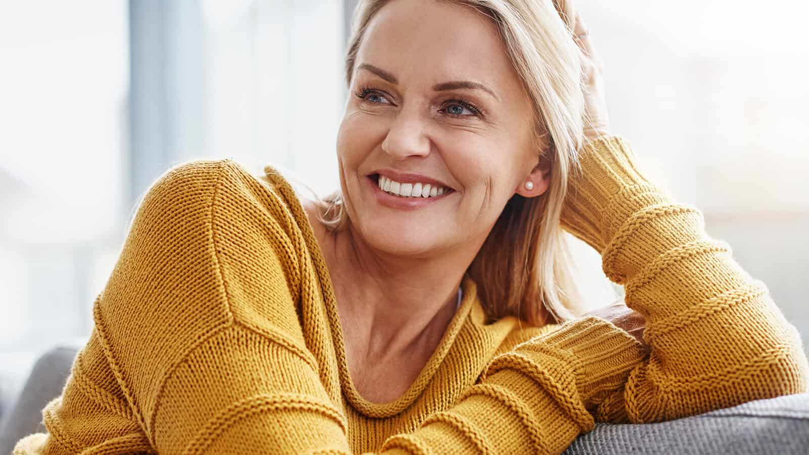 A smiling woman with blonde hair wearing a yellow sweater sits on a couch, resting her head on her hand and looking off to the side in a bright, cozy room.