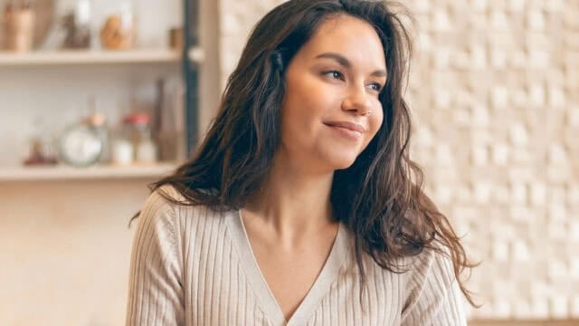 A woman with long brown hair and a beige top sits indoors, smiling softly and looking to the side. The background is softly lit with shelves and decorative items.