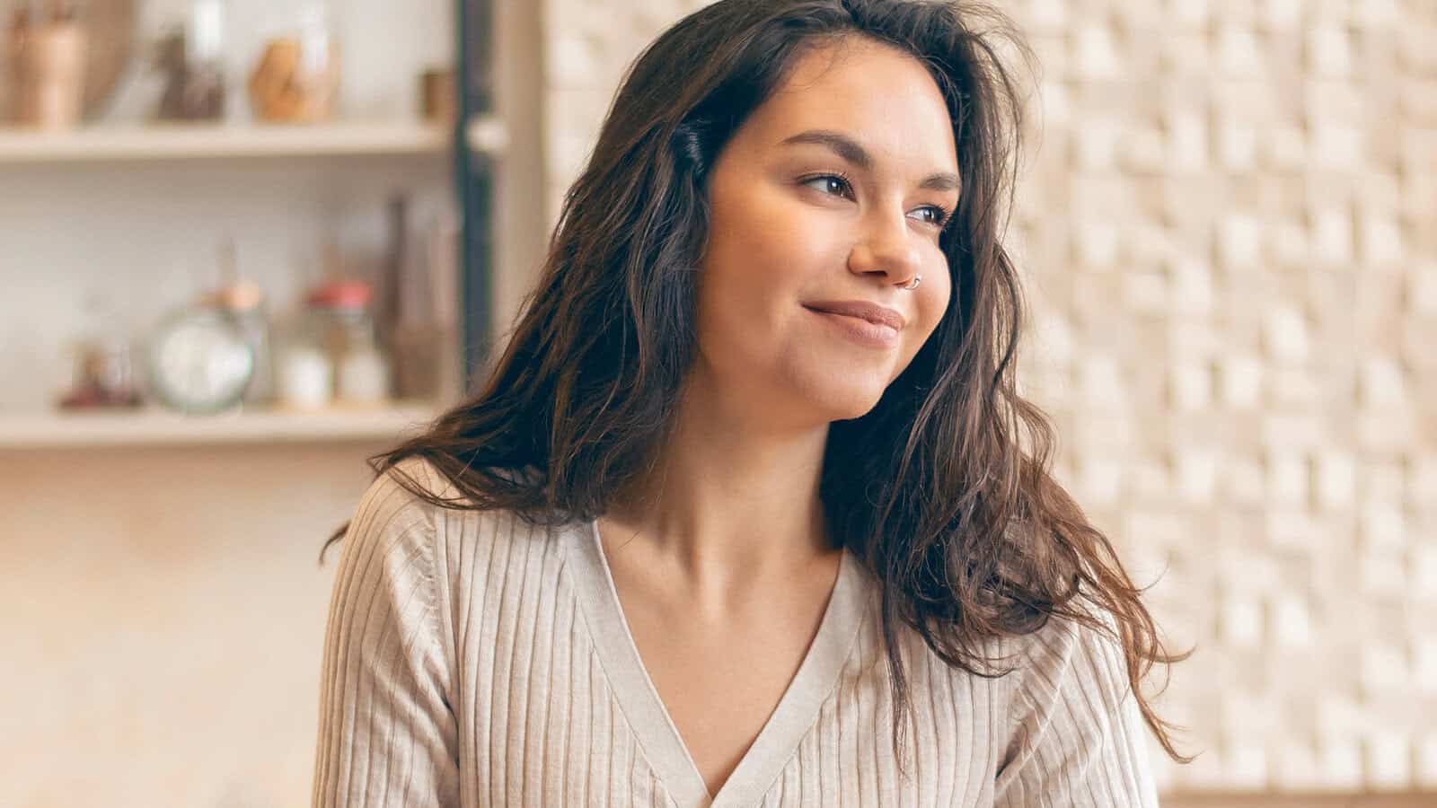 A woman with long brown hair and a beige top sits indoors, smiling softly and looking to the side. The background is softly lit with shelves and decorative items.