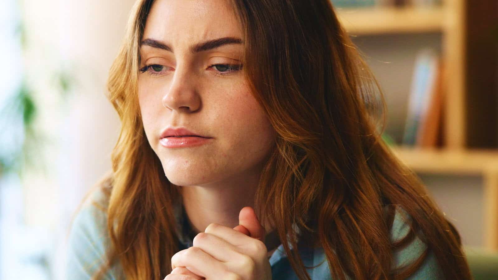 A young woman with long brown hair sits indoors, looking thoughtful and slightly concerned, with her hands clasped in front of her.
