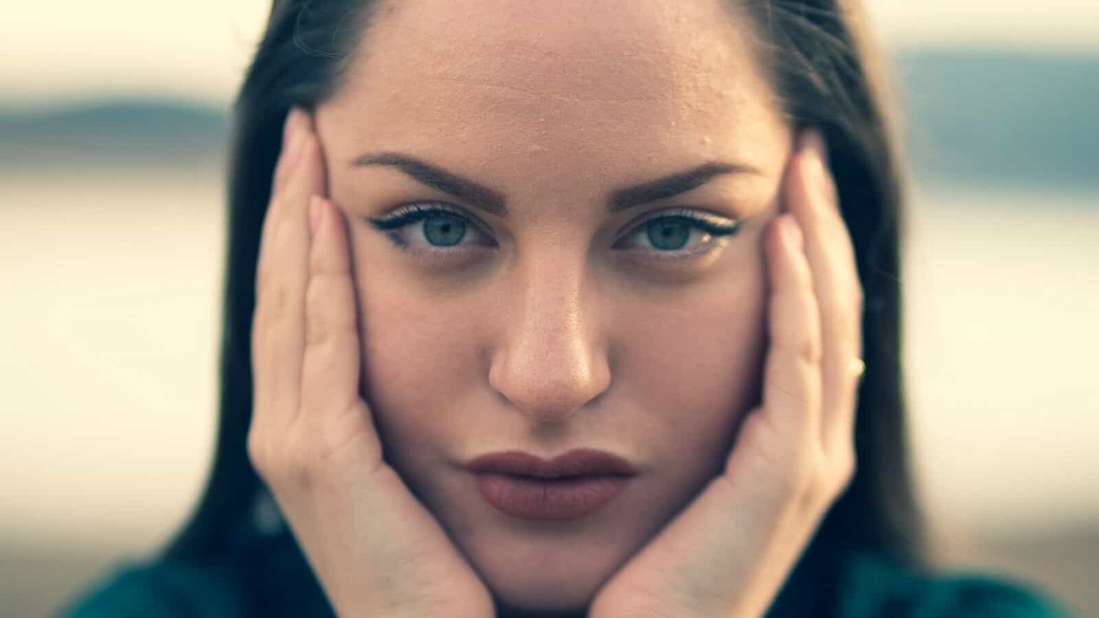 A woman with long dark hair holds her face in her hands, gazing intently at the camera. The background is softly blurred, focusing attention on her green eyes and neutral expression.