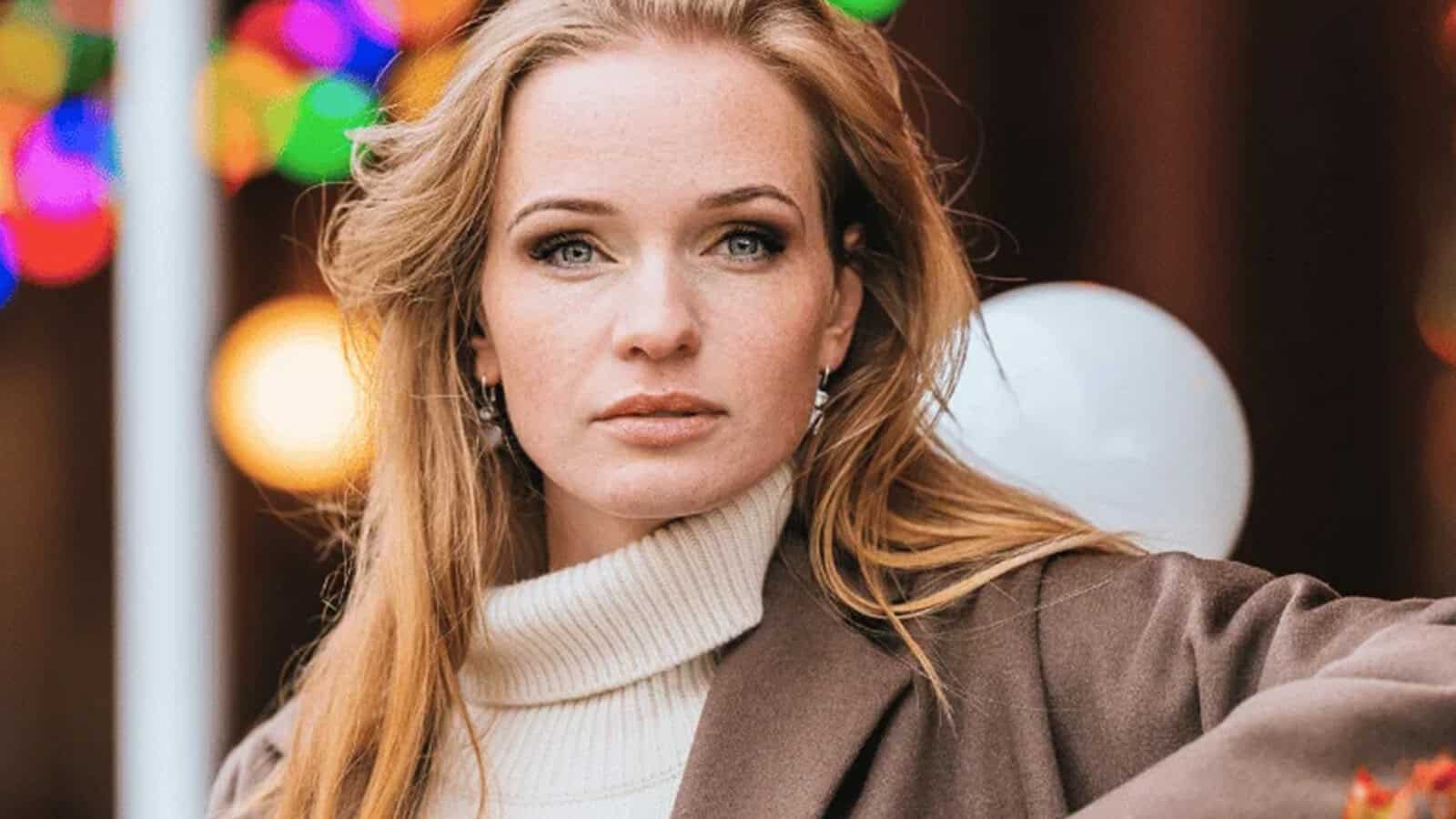 A woman with long blonde hair, wearing a white turtleneck sweater and brown coat, looks at the camera. Colorful lights and a blurred background are behind her.