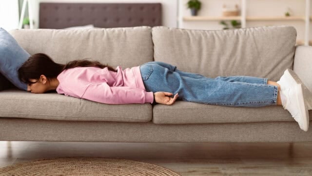 A person in a pink sweater and blue jeans lies face down on a gray couch, arms at their sides, with their head resting on a pillow.