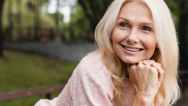 Smiling woman with long blonde hair wearing a light pink sweater, sitting outside on a bench, resting her chin on her hands, and looking to the side. Blurred background with green trees.