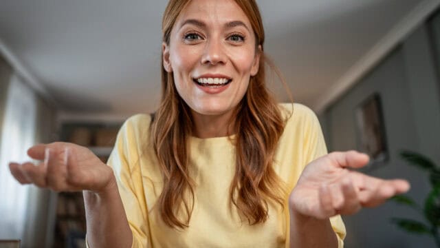 A woman with long red hair in a yellow shirt smiles and gestures with both hands, palms up, as if asking a question or expressing curiosity in a home setting.