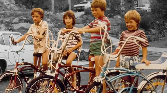 Four children in striped and patterned shirts sit on colorful vintage bikes with tall handlebars, smiling outdoors near parked cars and rocks, in a retro-style photo that beautifully captures the spirit of childhood during the 60s and 70s.