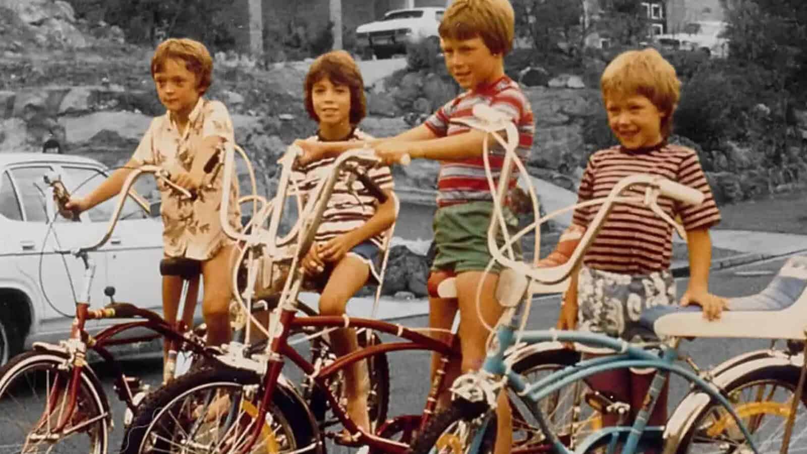 Four children in striped and patterned shirts sit on colorful vintage bikes with tall handlebars, smiling outdoors near parked cars and rocks, in a retro-style photo that beautifully captures the spirit of childhood during the 60s and 70s.
