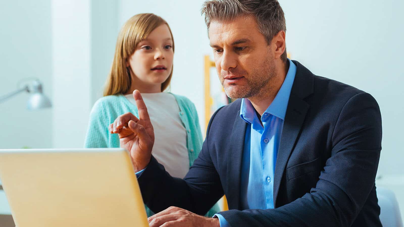 A man in a suit sits at a desk using a laptop, raising his finger as if making a point. A young girl stands behind him, looking at the screen, both appearing focused in a bright room.