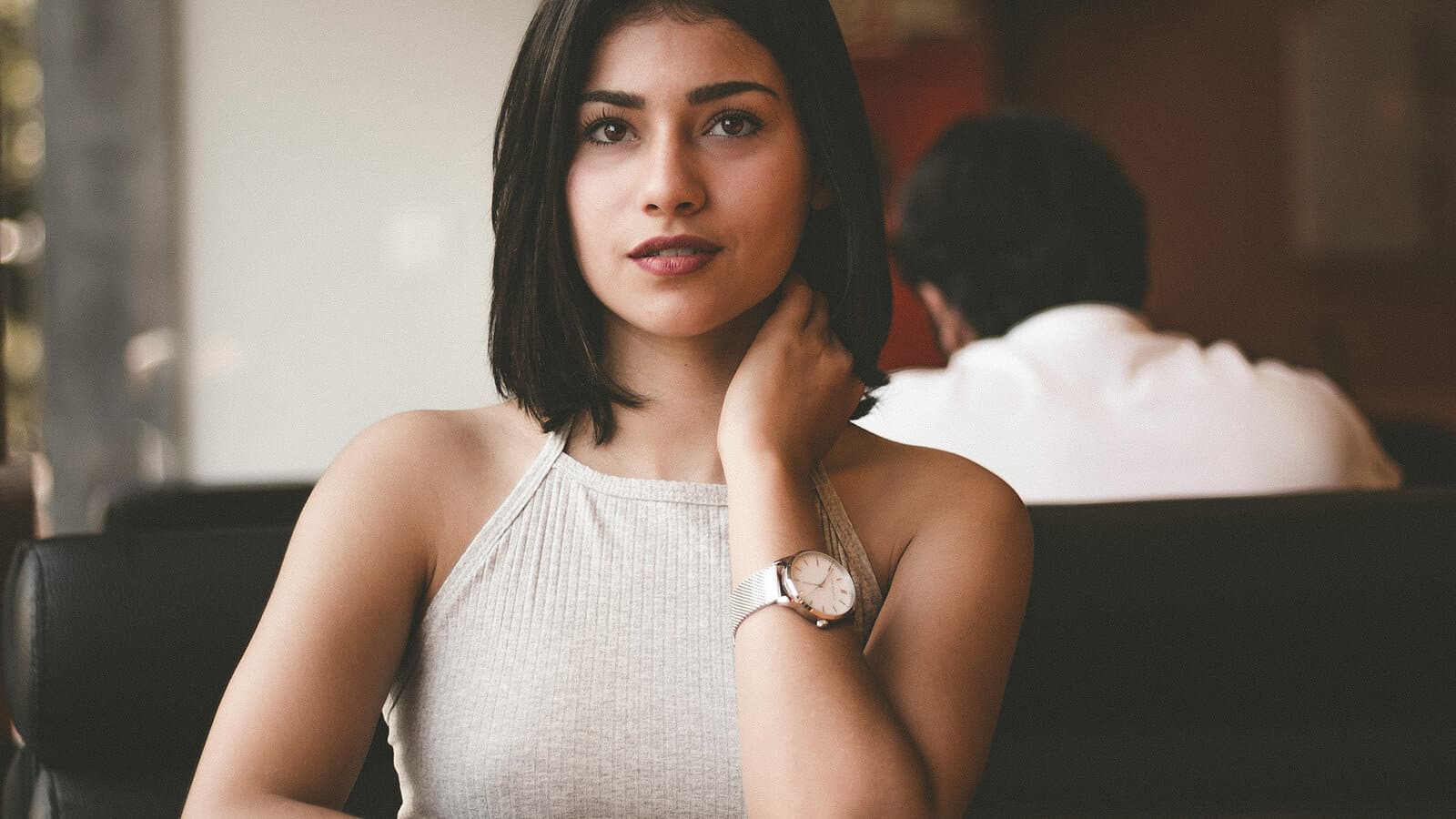 A woman with shoulder-length dark hair, wearing a sleeveless top and a wristwatch, sits on a black sofa, looking at the camera. A person with dark hair sits blurred in the background.