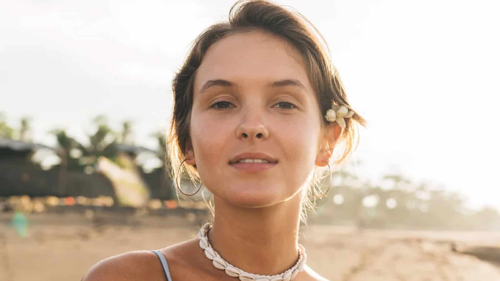 A young woman with short brown hair and a flower behind her ear stands on a beach, wearing a shell necklace and hoop earrings. The sun is shining and trees are visible in the blurred background.
