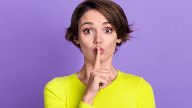 Woman with short brown hair wearing a bright yellow top holds her index finger to her lips, signaling shh against a solid purple background.