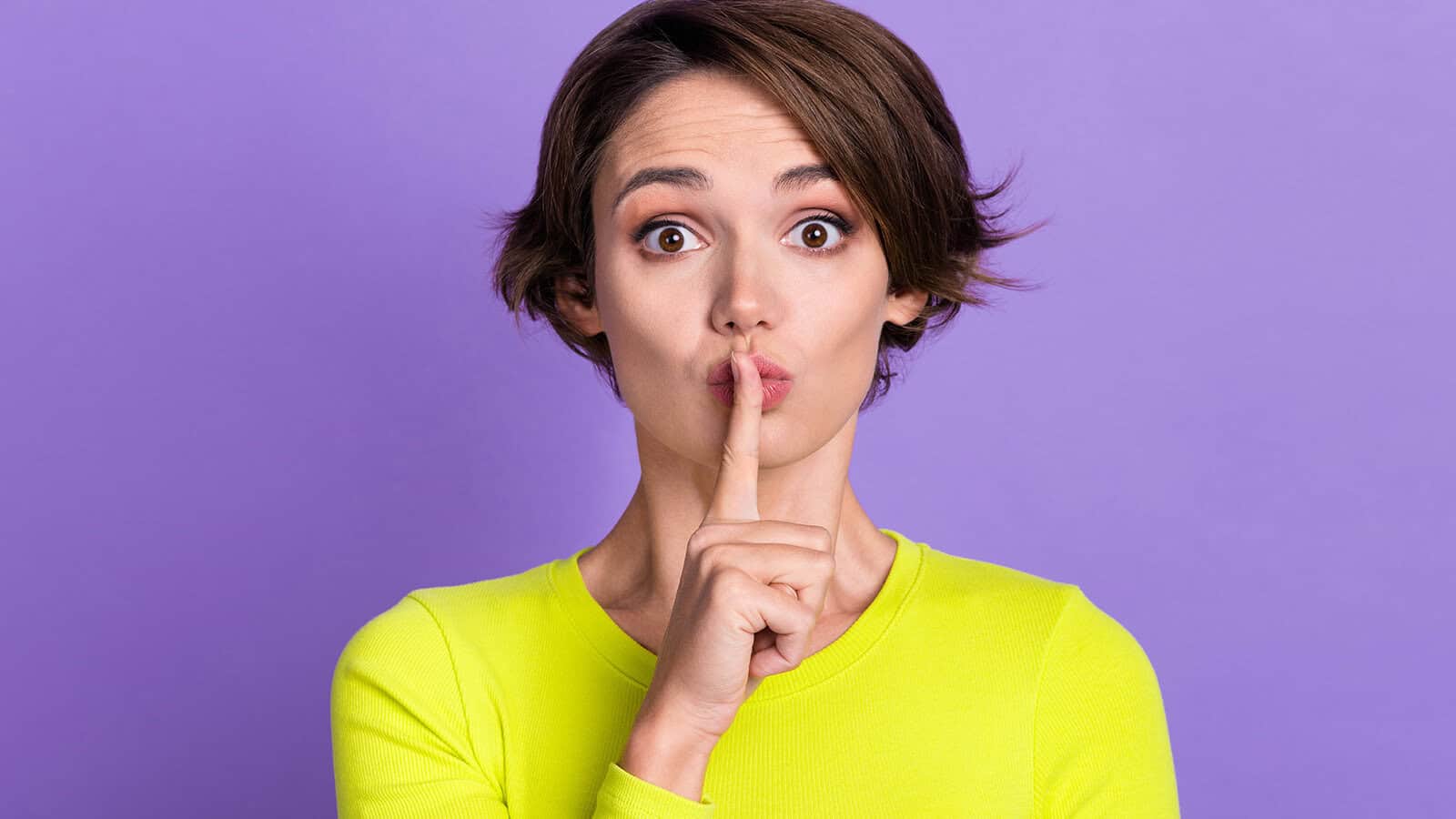 Woman with short brown hair wearing a bright yellow top holds her index finger to her lips, signaling shh against a solid purple background.