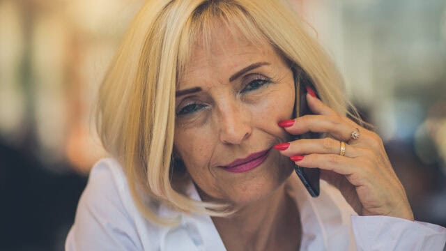 A middle-aged woman with blonde hair and pink lipstick holds a phone to her ear, looking directly at the camera with a slight smile. She is wearing a white shirt and has red painted nails.