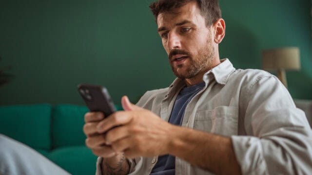 A man with short hair and a beard looks intently at his smartphone while sitting indoors on a teal couch, with a green wall and a lamp in the background.