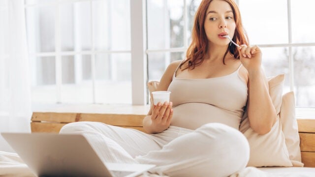 A pregnant woman sits cross-legged on a bed, eating yogurt with a spoon, and looking to the side. She wears light-colored clothes and has a laptop in front of her. Sunlight streams in through large windows behind her.