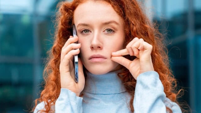 A woman with long red hair holds a smartphone to her ear while gently pinching her cheek with her other hand. She wears a light blue turtleneck and looks directly at the camera with a neutral expression.