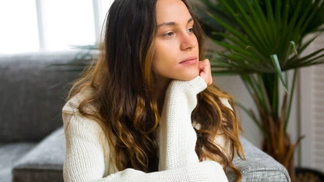 A woman with long brown hair, wearing a white sweater, sits on a couch with her chin resting on her hand, looking thoughtfully into the distance. Green plants and a window are in the background.