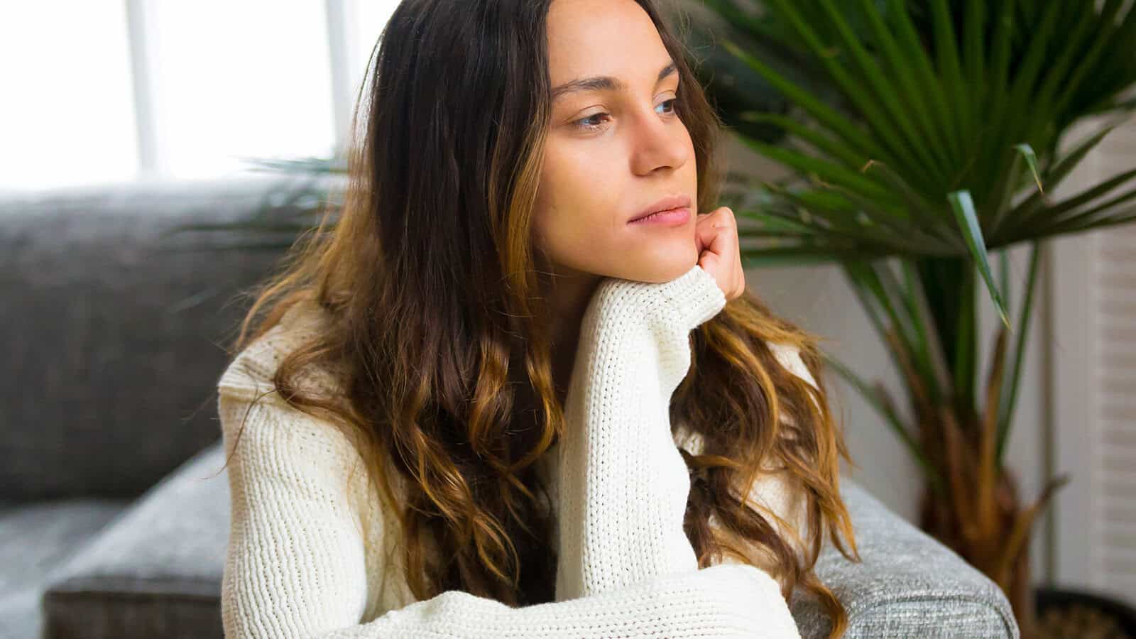 A woman with long brown hair, wearing a white sweater, sits on a couch with her chin resting on her hand, looking thoughtfully into the distance. Green plants and a window are in the background.