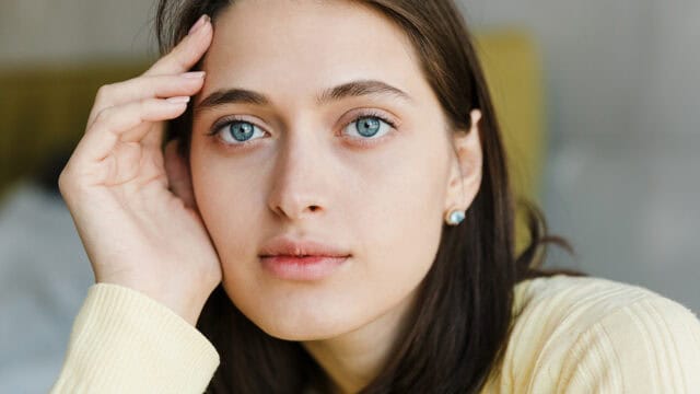 A young woman with light skin, blue eyes, and dark hair rests her head on her hand, wearing a yellow sweater and small earrings, looking thoughtfully at the camera.