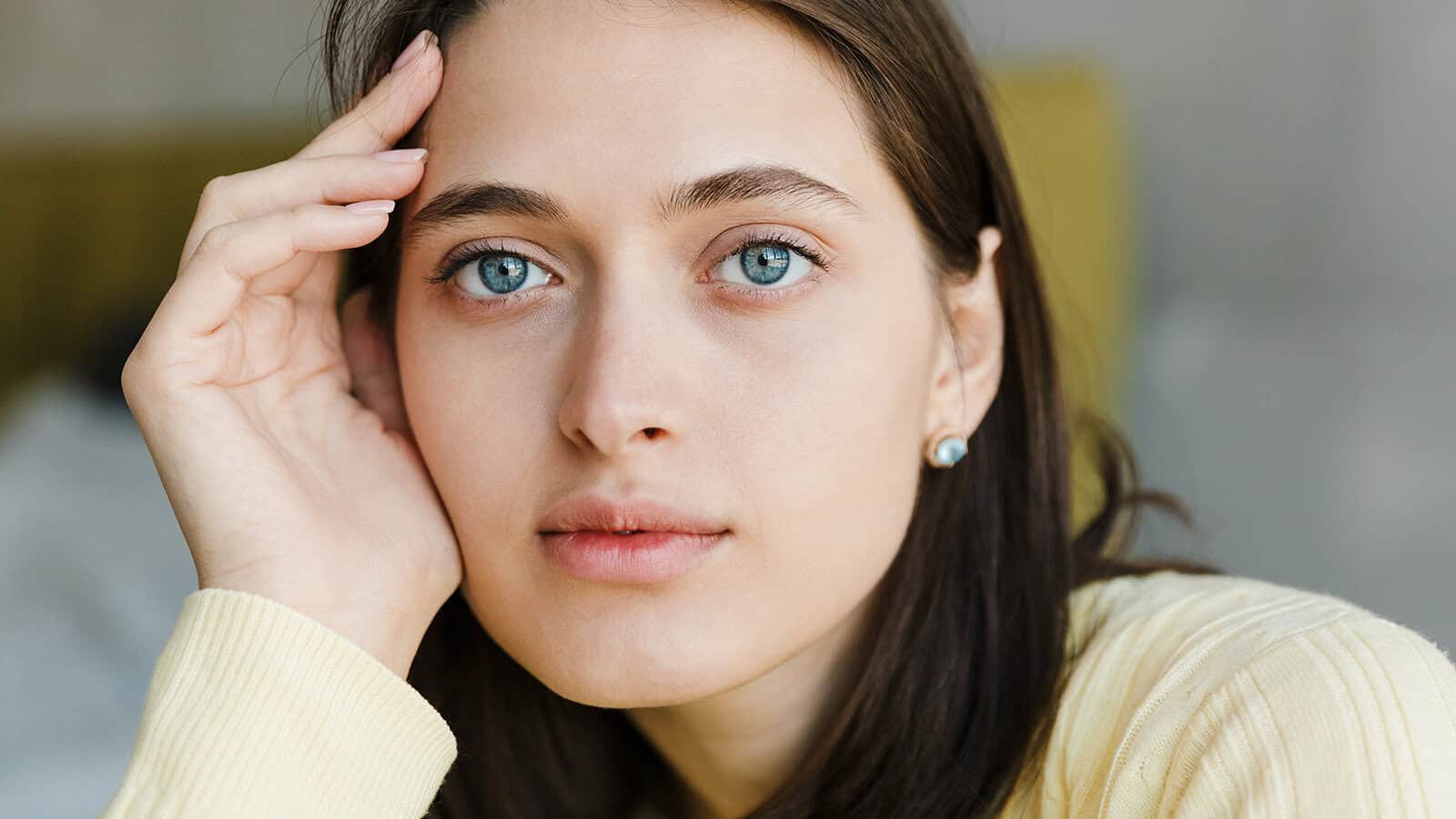 A young woman with light skin, blue eyes, and dark hair rests her head on her hand, wearing a yellow sweater and small earrings, looking thoughtfully at the camera.
