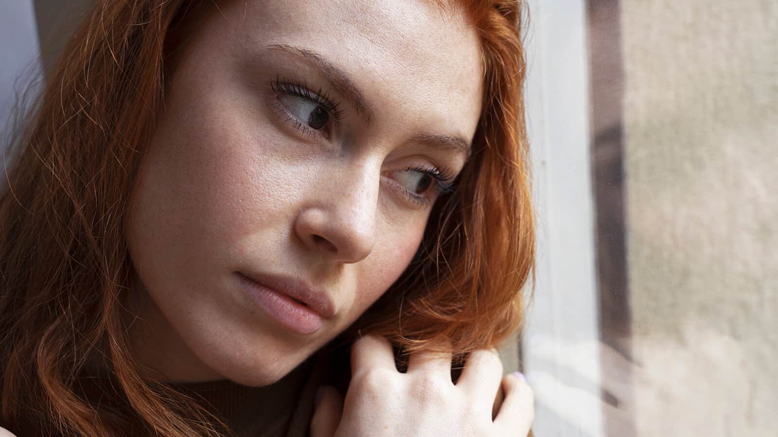 A woman with red hair gazes out a window, resting her chin on her hand, appearing thoughtful and pensive.